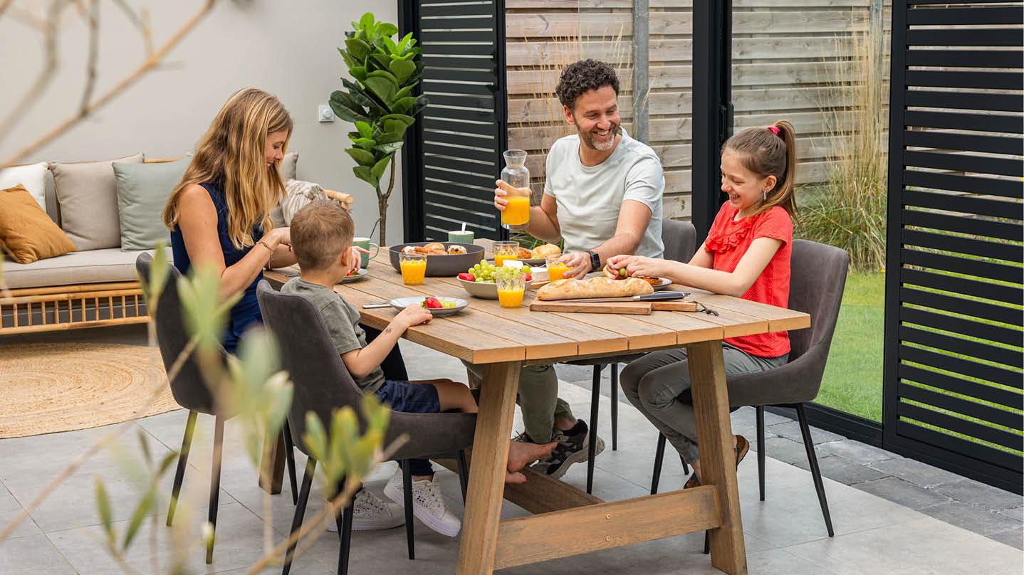 Familie geniet van een zonnig ontbijt, in een stijlvolle Gumax® terrasoverkapping Een glimlachende familie geniet samen van het ontbijt op een Gumax® terrasoverkapping met Shading Panels.